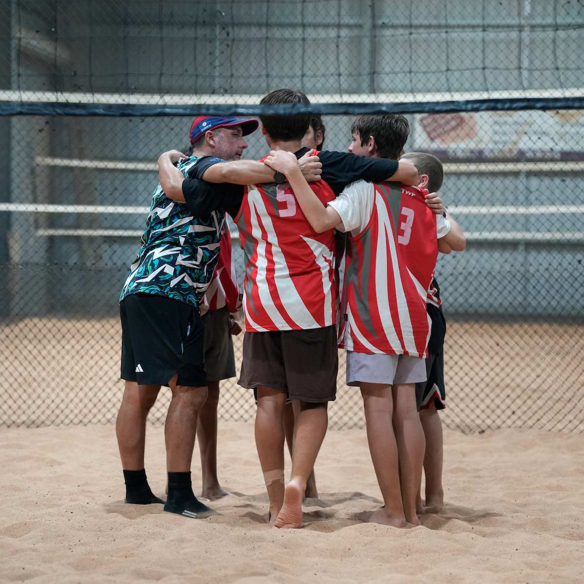 Coach and young players huddle together on a sandy sports court.